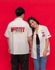 Two models wearing matching off-white racing-style short-sleeve shirts with retro patches and graphics. The man shows the back graphic reading 'MICKEY Racing', while the woman shows the front chest patches.