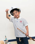Child wearing polo tee in stripe sailor mickey and shorts, holding a toy boat and oar, with a white wall background.