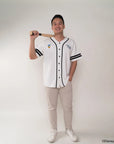 Person wearing a white baseball jersey with black accents, holding a bat on a plain background.