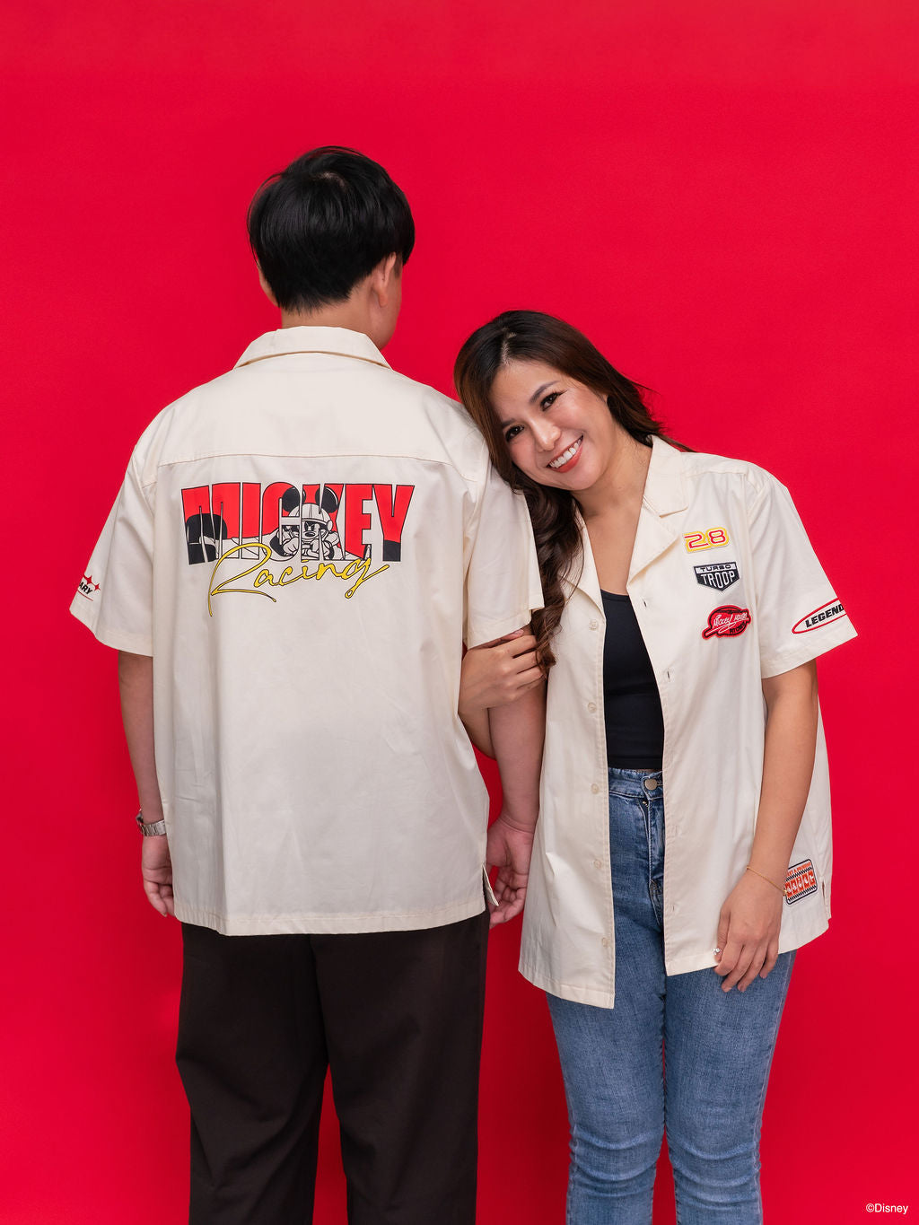 Two models wearing matching off-white racing-style short-sleeve shirts with retro patches and graphics. The man shows the back graphic reading 'MICKEY Racing', while the woman shows the front chest patches.
