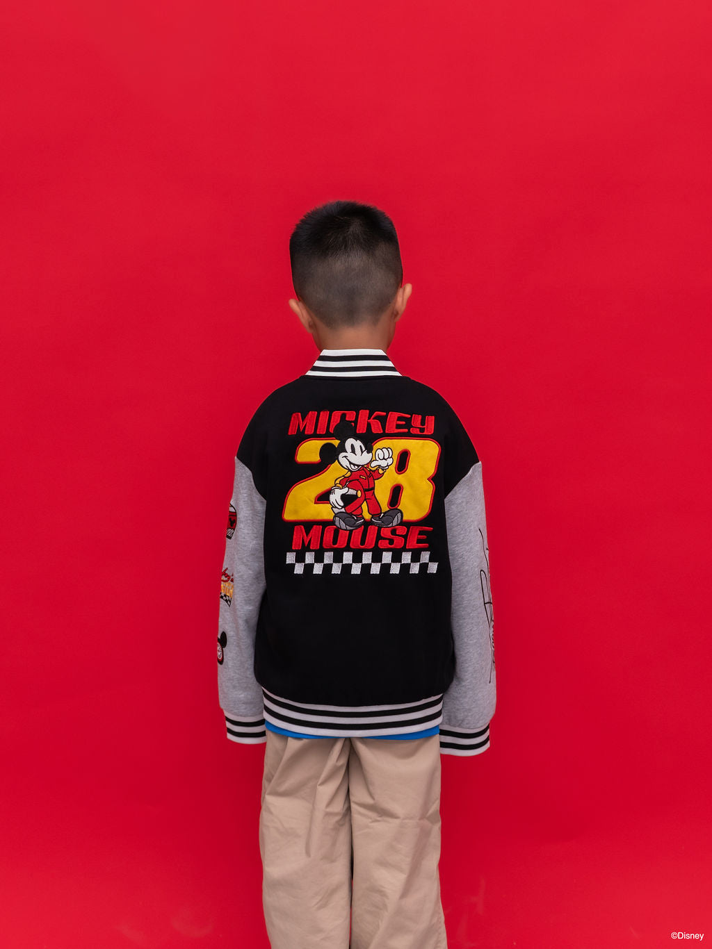 A child viewed from behind wearing a black and gray Mickey 28 Bomber jacket with a large Mickey Mouse '28' graphic on the back, standing against a solid red background.