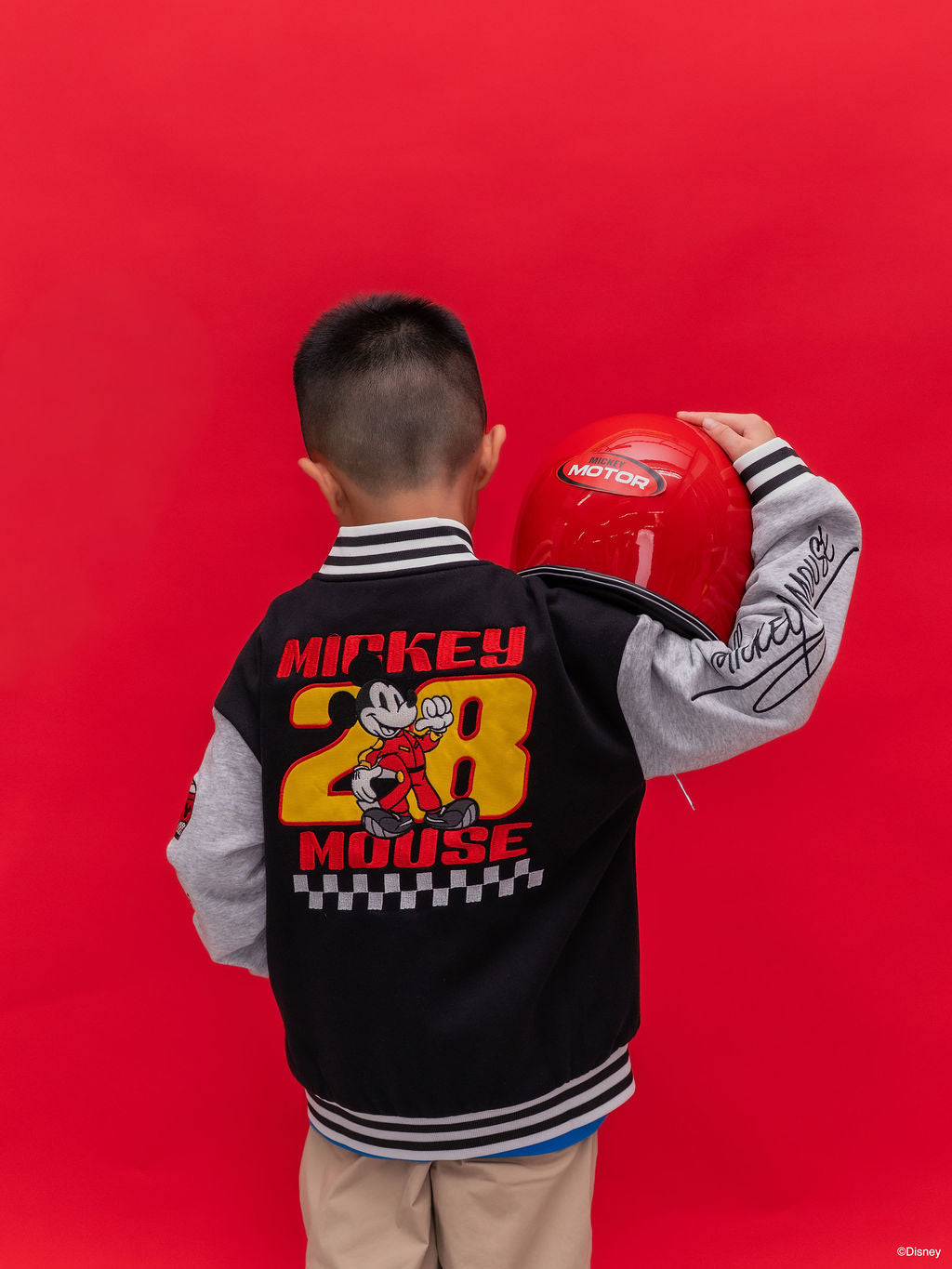A child shown from the back wearing a black and gray Bomber Jacket - Mickey 28 varsity jacket with a large Mickey Mouse embroidery on the back.