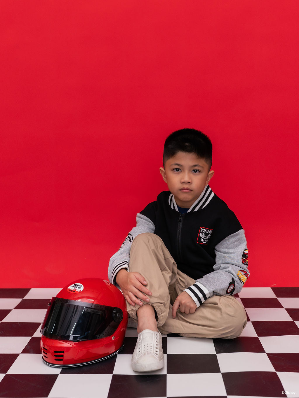 A young boy wear a black and gray Bomber Jacket - Mickey 28 varsity jacket and Native shoes next to a red full-face helmet.
