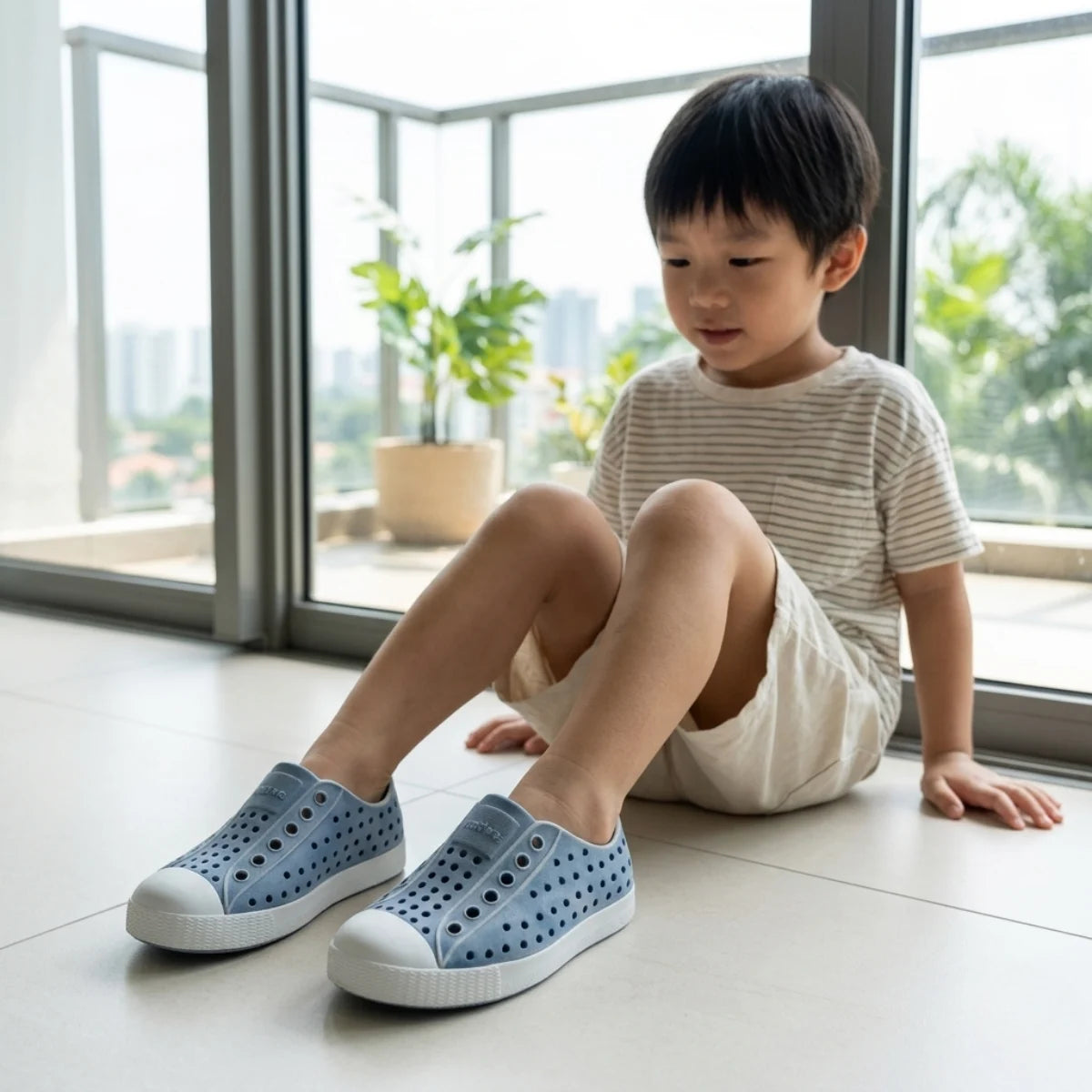 Child wearing blue shoes sitting on a floor with large windows in the background