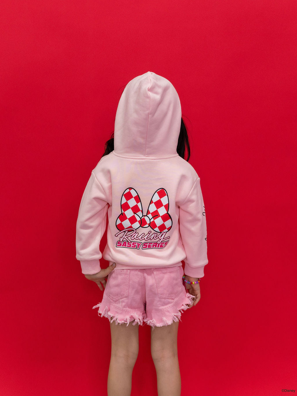 A child wearing a Minnie Mouse pink hoodie with a large red and white checkered bow graphic and 'Racing Sassy Series' text on the back. 