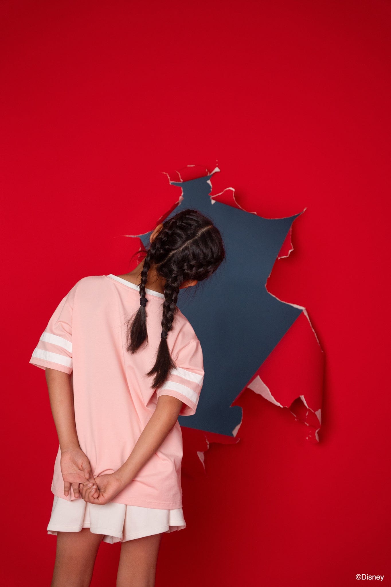 Girl in Stitch tee shirt looking through hole in red wall.