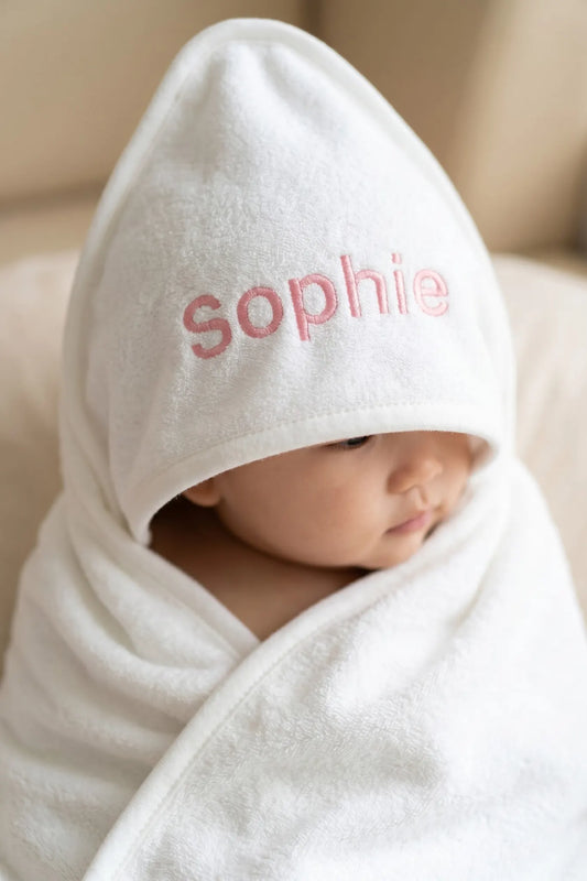 Baby wrapped in a white hooded towel with 'sophie' embroidered on it, sitting on a beige surface.