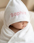 Baby wrapped in a white hooded towel with 'sophie' embroidered on it, sitting on a beige surface.