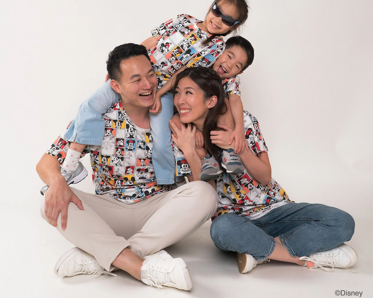 A family of four wearing matching Disney Mickey Mouse-themed short-sleeve t-shirts. The shirts feature a colorful collage print of Mickey Mouse and other Disney characters.